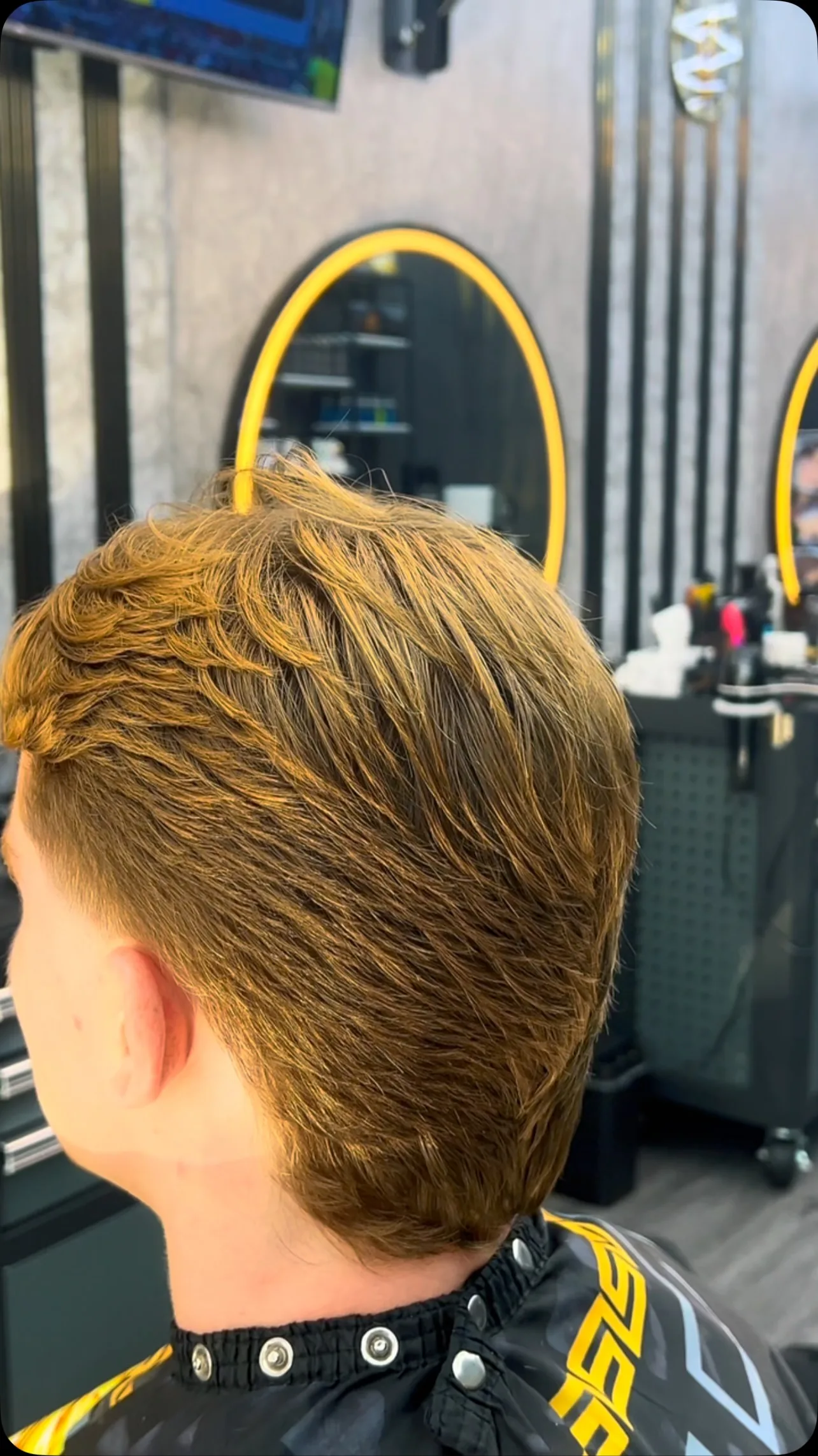 Back of head, clean mid-taper on wavy brown hair, round ring mirror and tool trolley.
