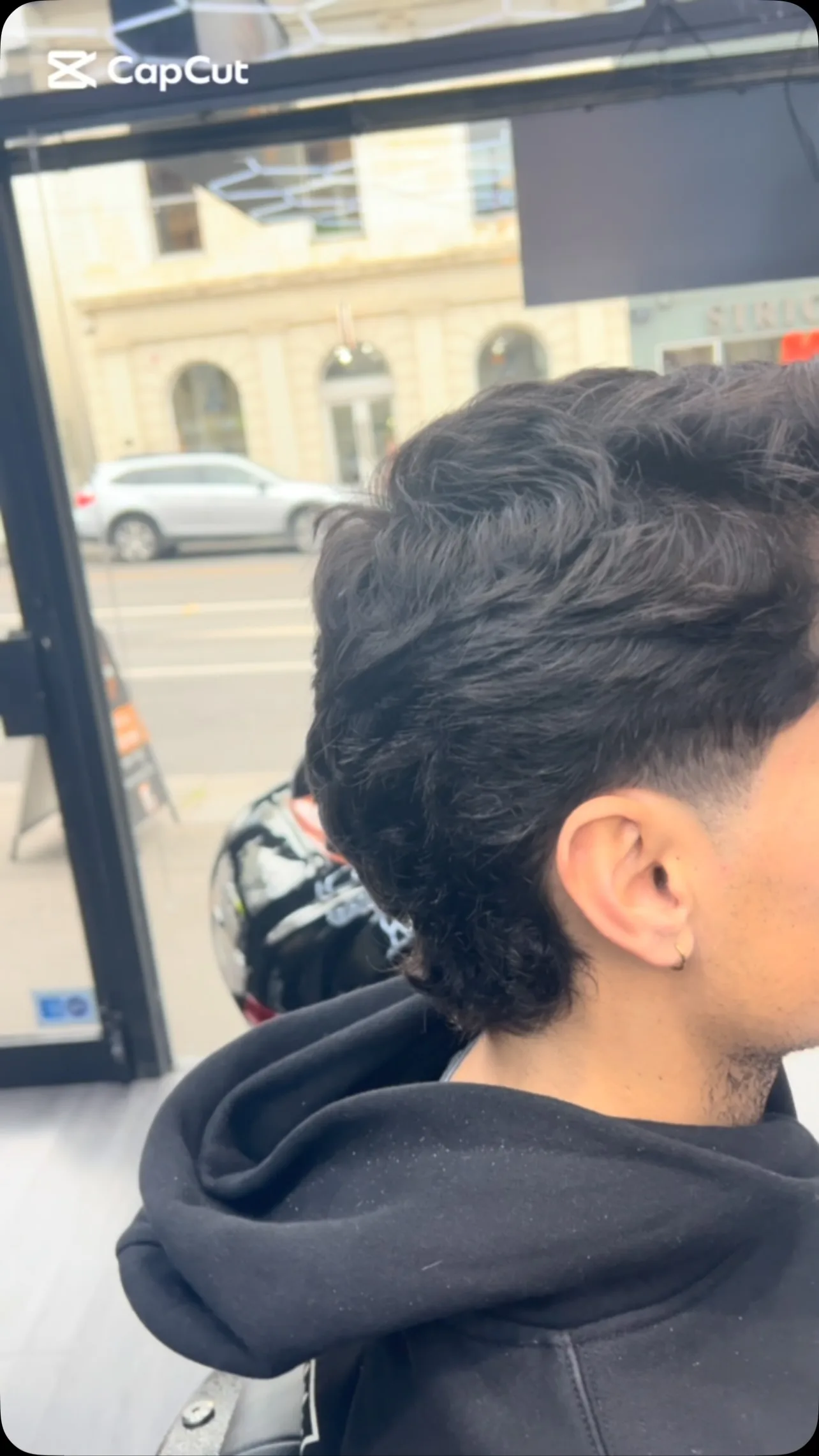 Rear three-quarter, curly black mullet, hoodie, storefront window and parked car outside.