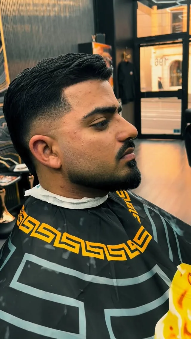 Low skin fade blended into full beard — Versace Greek-key cape, Bridge Road reflection