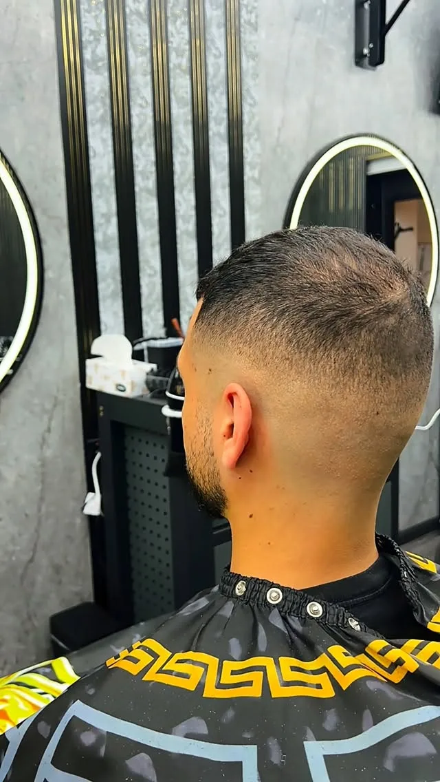 Rear, ginger buzz cut with high skin fade, neck tattoo, white and black-gold cape.