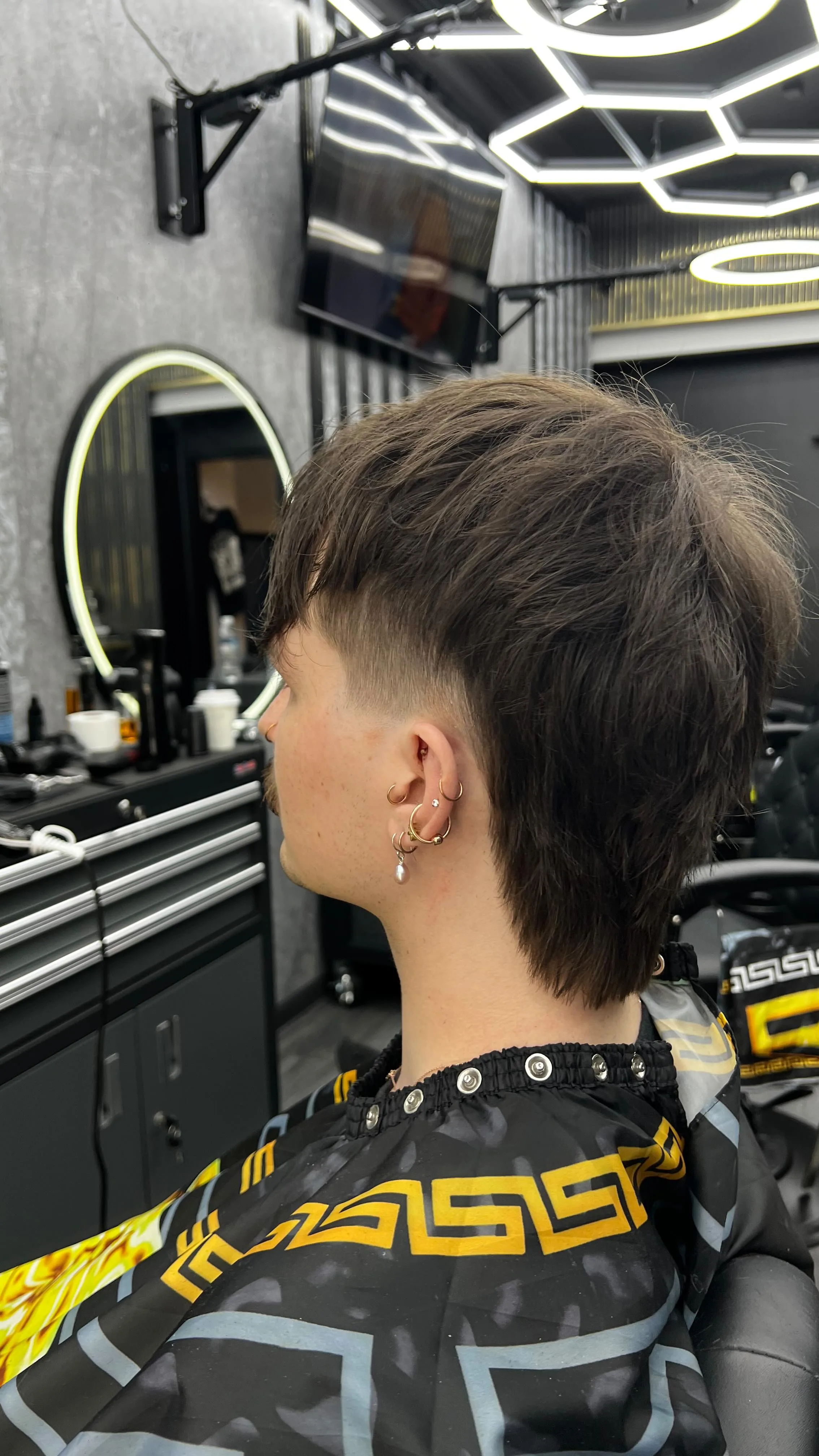 Rear three-quarter, fresh textured mullet and taper, Versace black and gold Greek-key cape, honeycomb LED ceiling above.